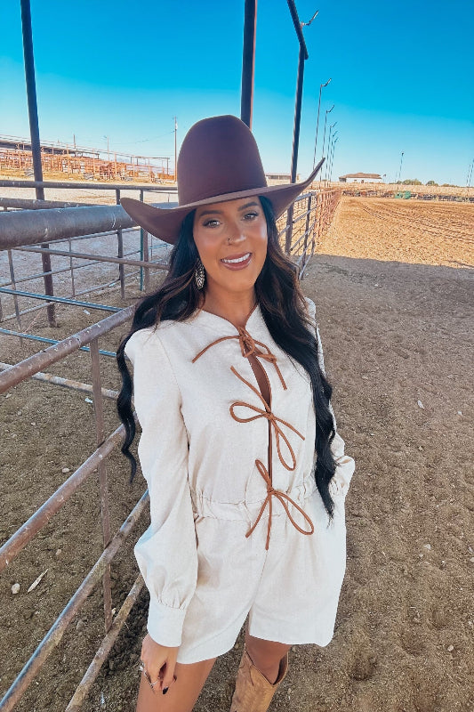 Woman wearing a white outfit with brown accents and a brown hat in an outdoor setting.