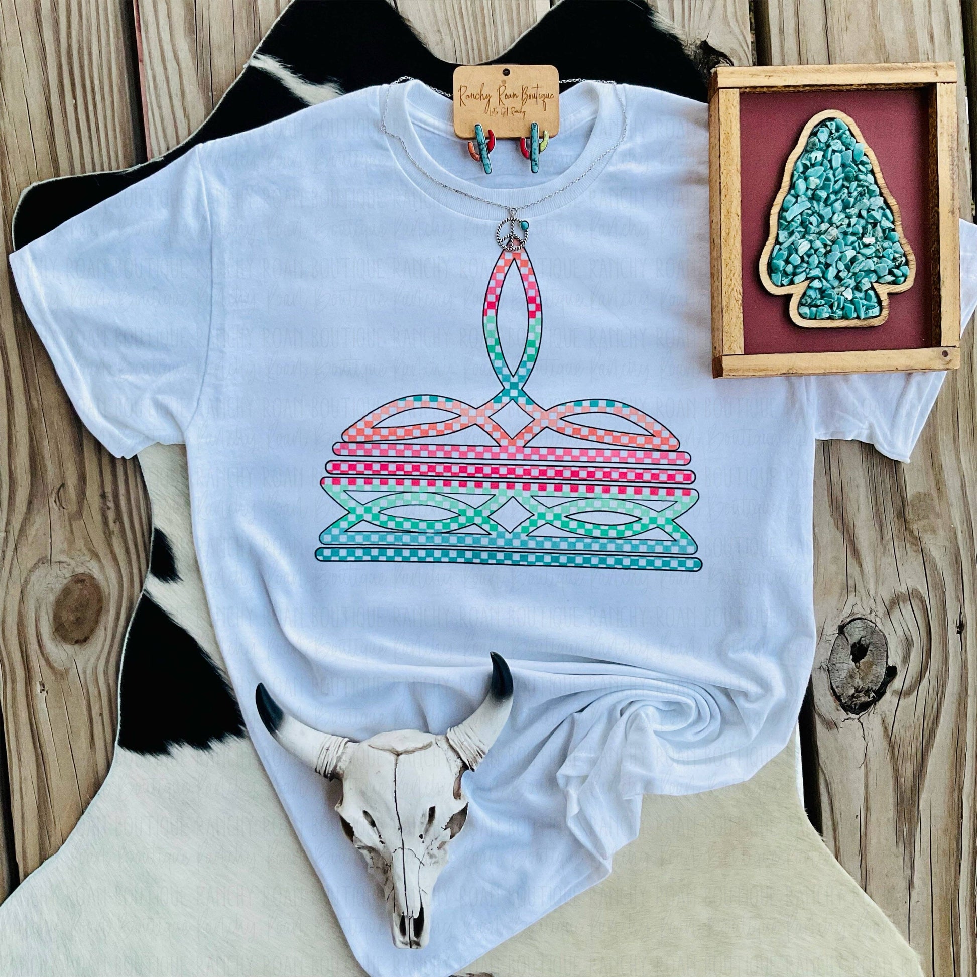 White t-shirt with a colorful graphic design on a wooden surface with a cow skull and framed artwork.