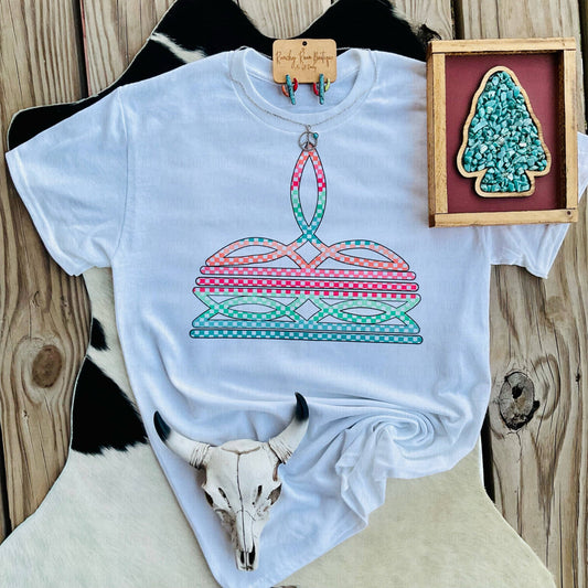 White t-shirt with a colorful graphic design on a wooden surface with a cow skull and framed artwork.