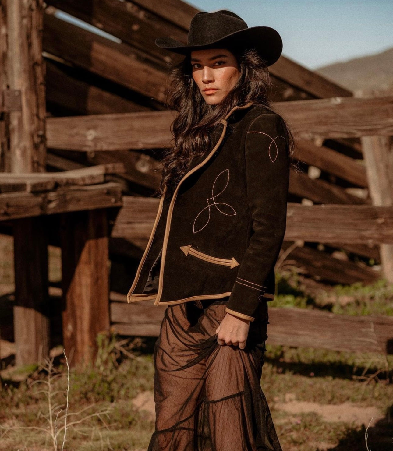 Woman in a cowboy hat and embroidered jacket standing in front of wooden structures.
