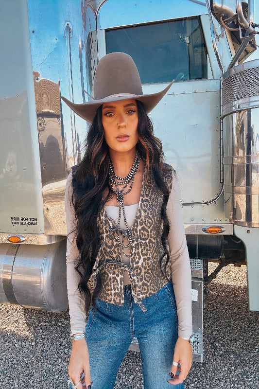 Woman wearing a cowboy hat, leopard print vest, and blue jeans standing in front of a metallic trailer.