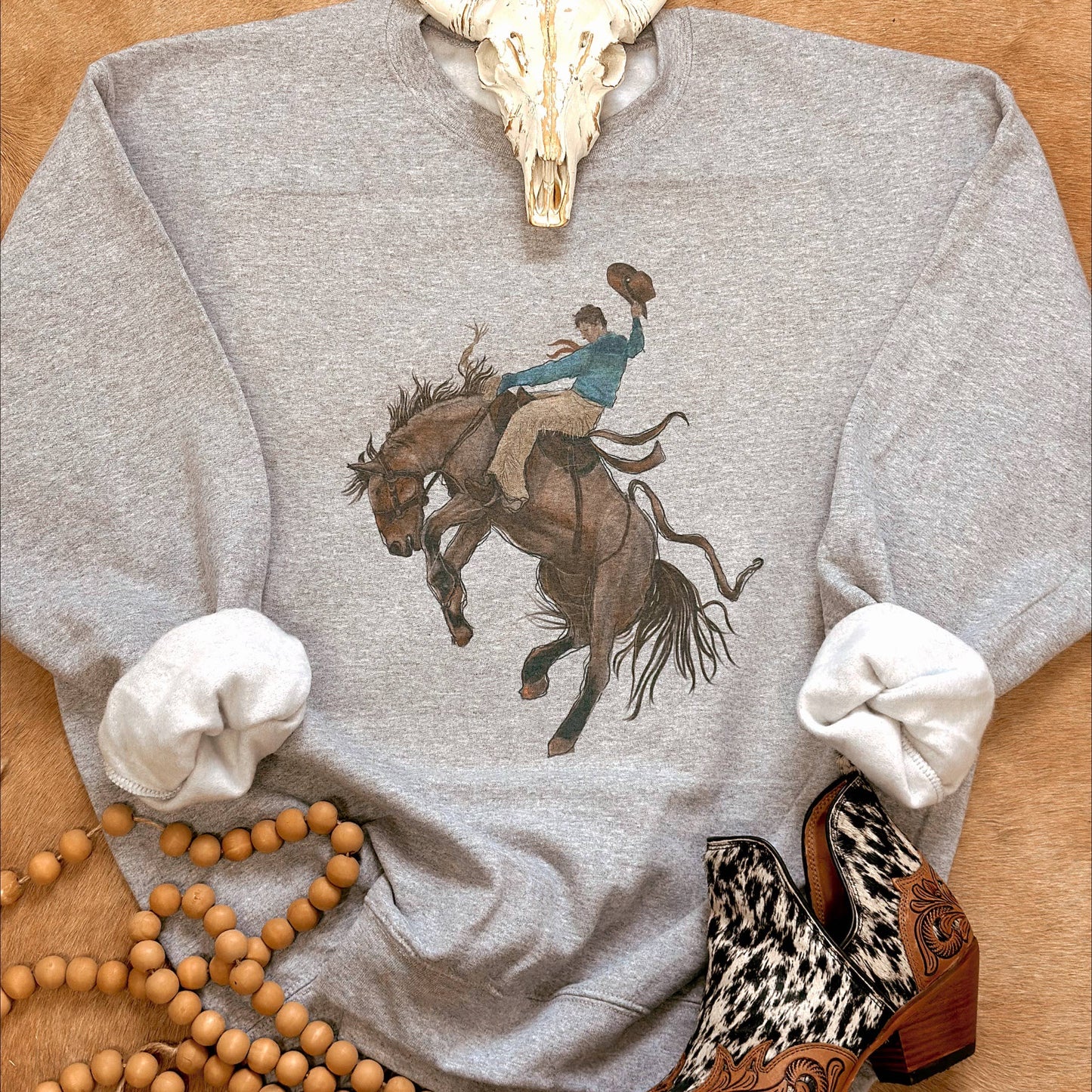 Gray sweatshirt with a graphic of a person riding a horse, surrounded by decorative items on a wooden surface.