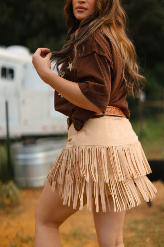 Woman wearing a brown top and beige fringed skirt outdoors.