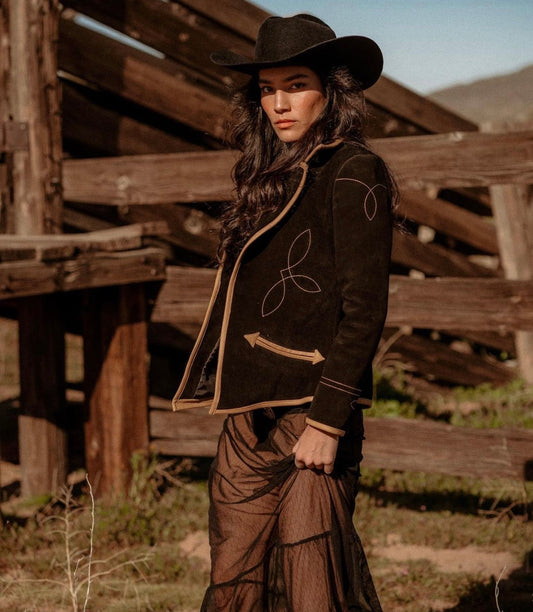 Woman in a cowboy hat and embroidered jacket standing in front of wooden structures.
