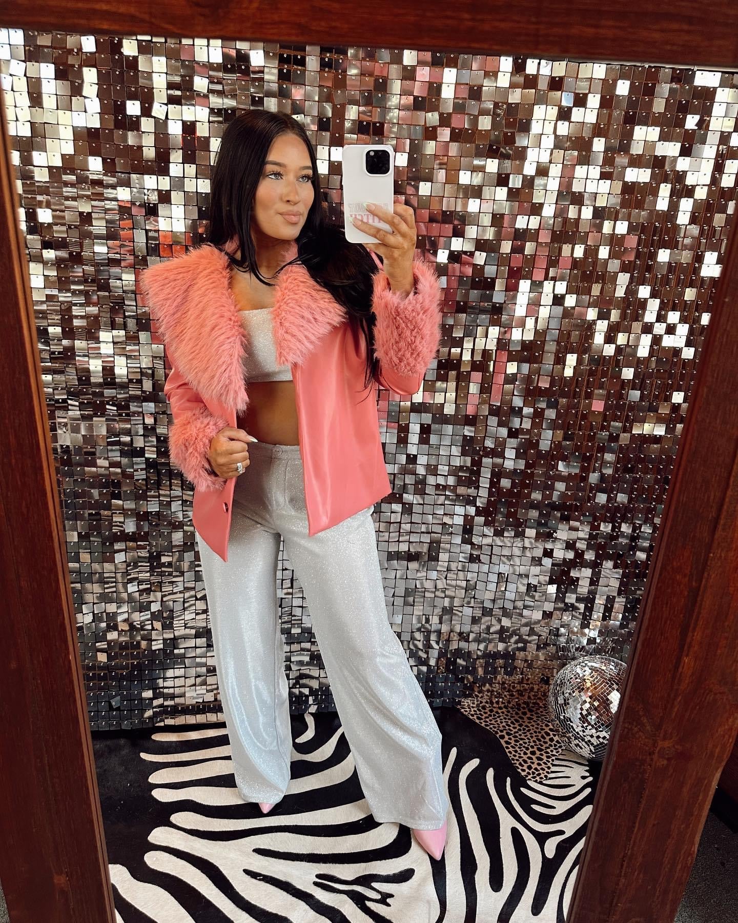 Person wearing a pink fur coat and white pants standing in front of a mirror with a decorative wall.